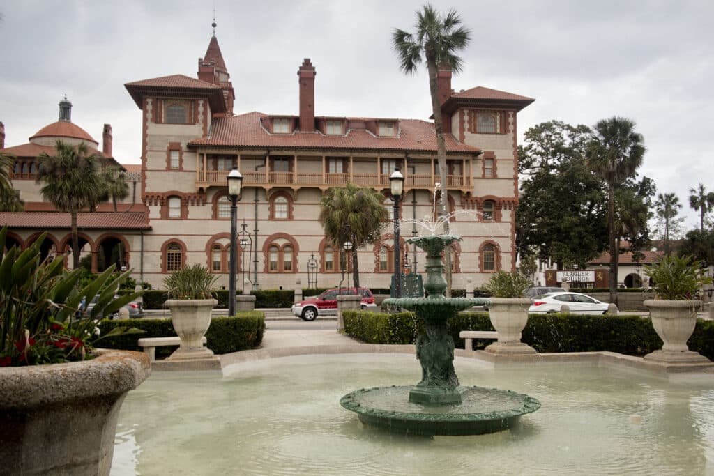 Um edifício histórico com telhado vermelho e torres atrás de uma fonte verde, cercado por palmeiras e carros estacionados.
