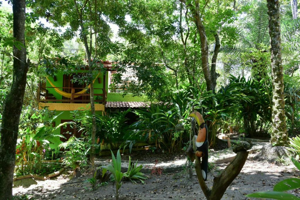 Um edifício verde da Casa Mestiça com uma varanda é cercado por plantas e árvores densas; uma estátua de tucano está em primeiro plano.