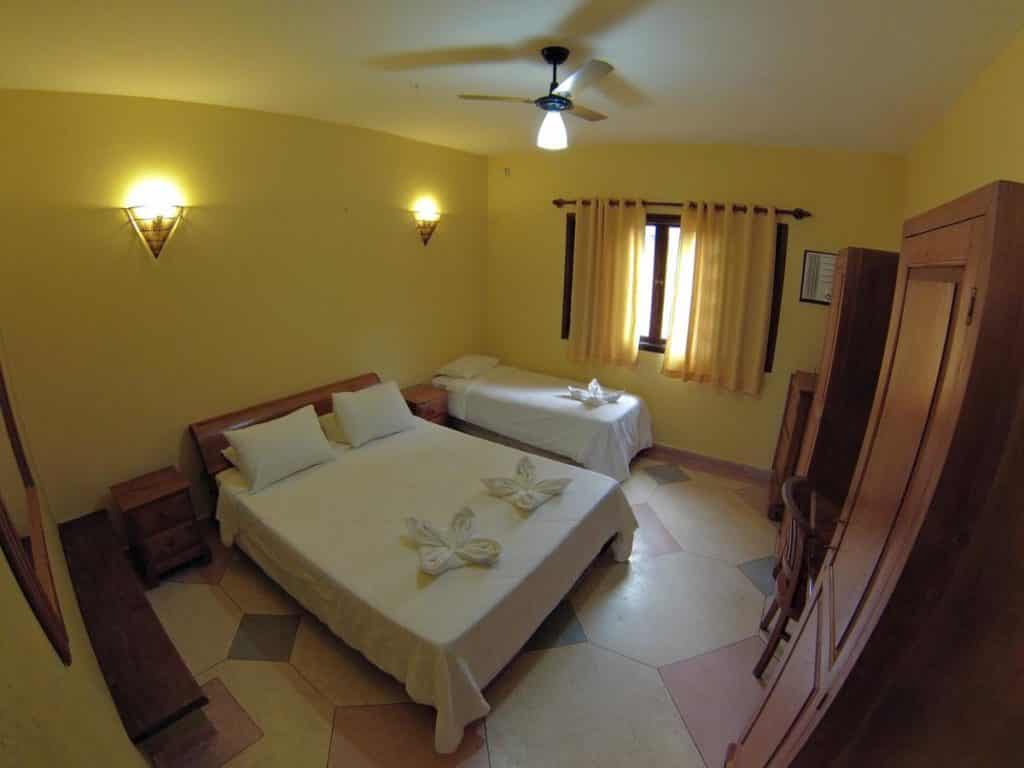 Um quarto do Hotel Pousada dos Condes com paredes amarelas, uma cama de casal, uma cama de solteiro, um guarda-roupa, um ventilador de teto e uma janela com cortinas.