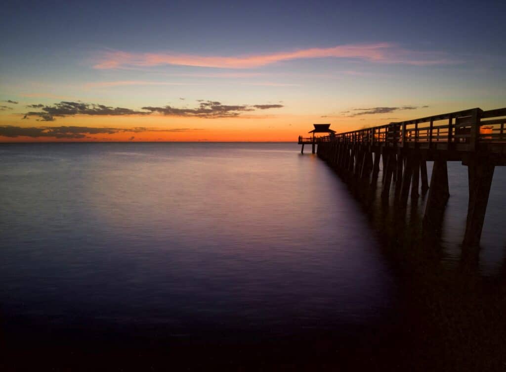 Pôr do sol sobre o oceano com um píer de madeira vazio se estendendo até a água, com um céu laranja e roxo ao fundo.