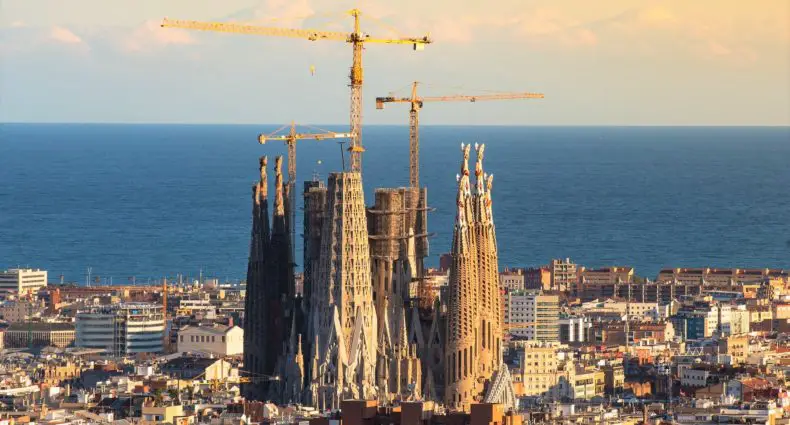 Vista aérea da Sagrada Família em Barcelona com guindastes de construção, paisagem urbana e mar ao fundo. A Igreja é uma das principais escolhas de o que fazer em Barcelona.