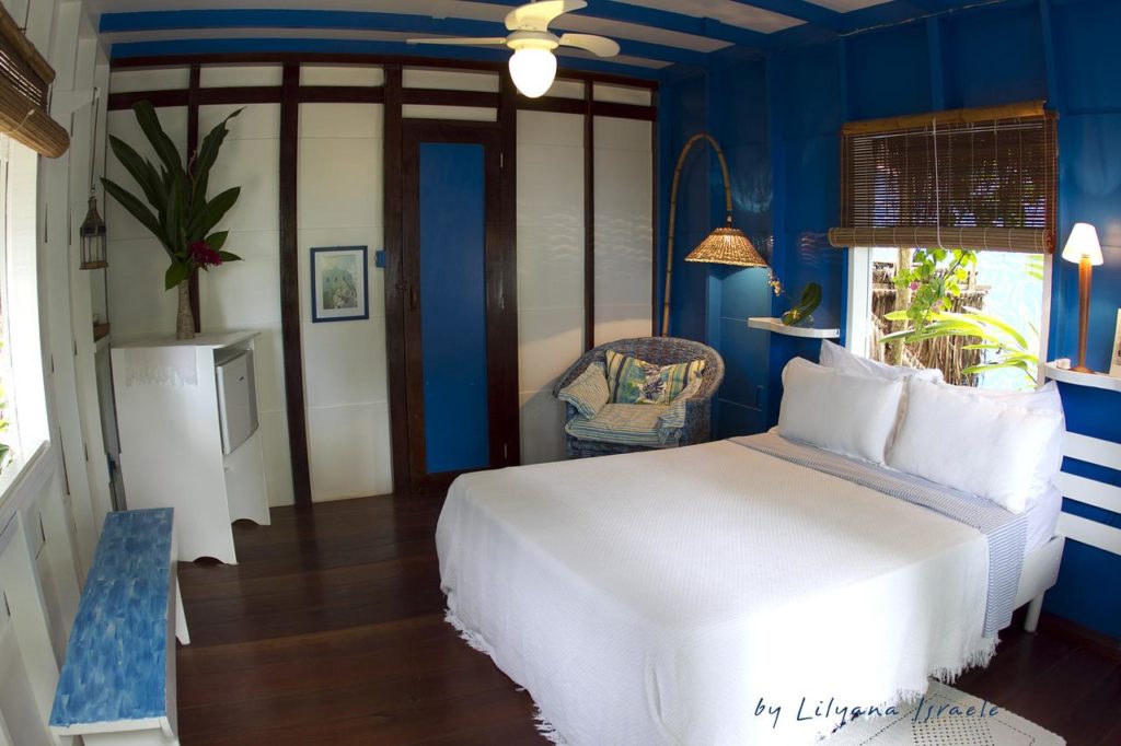 Um quarto pequeno e bem iluminado da Pousada Vila Sereia com paredes azuis e brancas, uma cama de casal, cadeira, ventilador de teto e janela com luz natural.