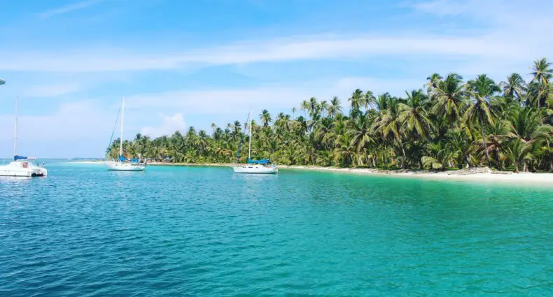 Três veleiros flutuam perto de uma costa tropical ladeada por palmeiras sob um céu azul claro em San Blas, no Panamá, para representar onde ficar em San Blas.