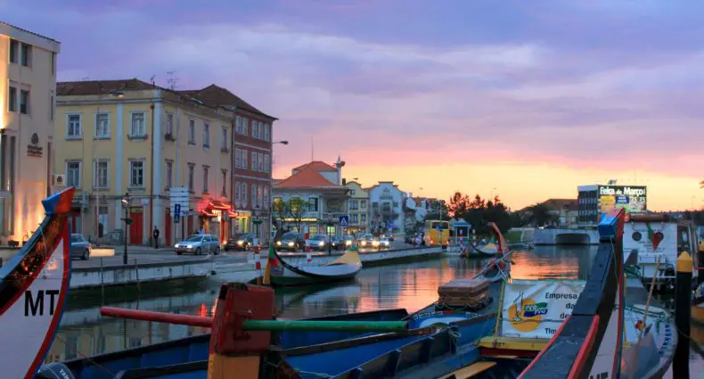 Entardecer na cidade de Aveiro, com prédios próximos ao rio navegado por barcos. Foto de Nmmacedo via Wikimedia.