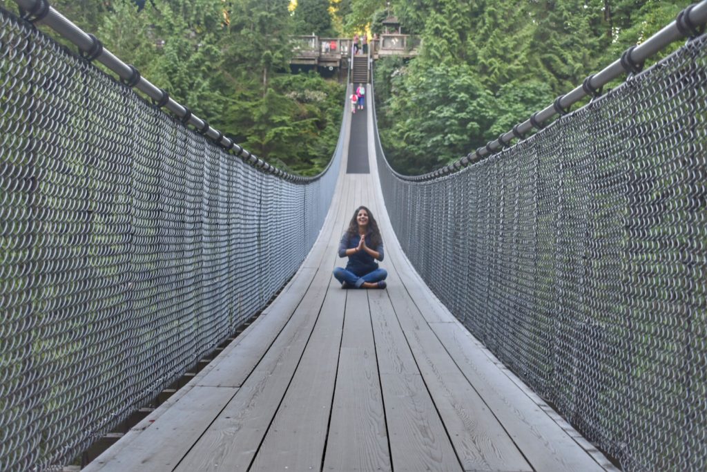 Uma de nossas colunistas está sentada de pernas cruzadas em uma ponte pênsil cercada por árvores verdes exuberantes, com as mãos em uma pose meditativa. Imagem para ilustrar post sobre o que fazer em Vancouver.