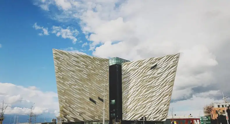 Vista de fora do Titanic Belfast. O Edifício é moderno, em formato de navio, com fachadas angulares e metálicas e painéis externos padronizados sob um céu parcialmente nublado, situado em um gramado. A foto está no post de o que fazer em Belfast.