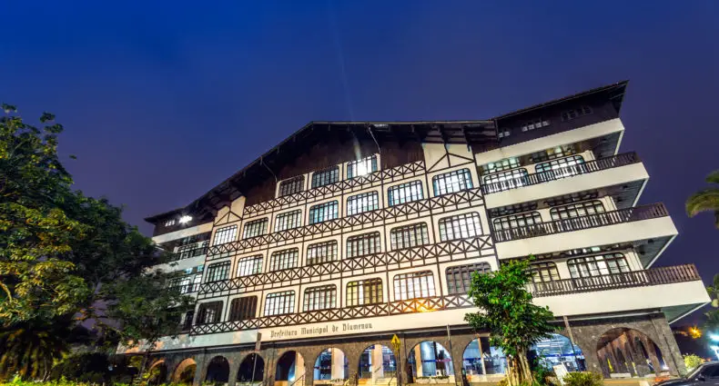 Um grande edifício de vários andares com estrutura de madeira, iluminado à noite, com árvores e uma área de paralelepípedos na frente.