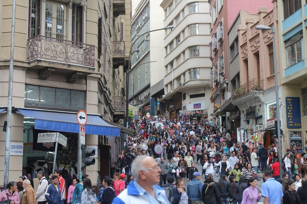 Vista de esquina da Rua 25 de Março, um dos pontos turísticos de SP, com multidão de pessoas em rua inclinada. Foto de C. Araujo26 via Wikimedia