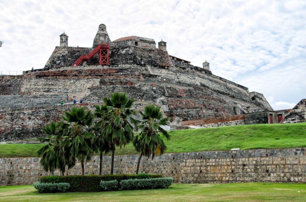 A imagem mostra o Castillo San Felipe de Barajas, em Cartagena, uma fortaleza histórica imponente feita de pedra. A estrutura tem várias camadas de muralhas que se elevam em direção ao topo, onde há pequenas torres e uma escada vermelha visível. Na base da fortaleza, há uma área verde bem cuidada com palmeiras e arbustos. A parede de pedra no nível mais baixo é robusta e antiga, adicionando uma sensação de solidez e durabilidade ao local. O céu está parcialmente nublado, proporcionando uma luz suave sobre a cena.
