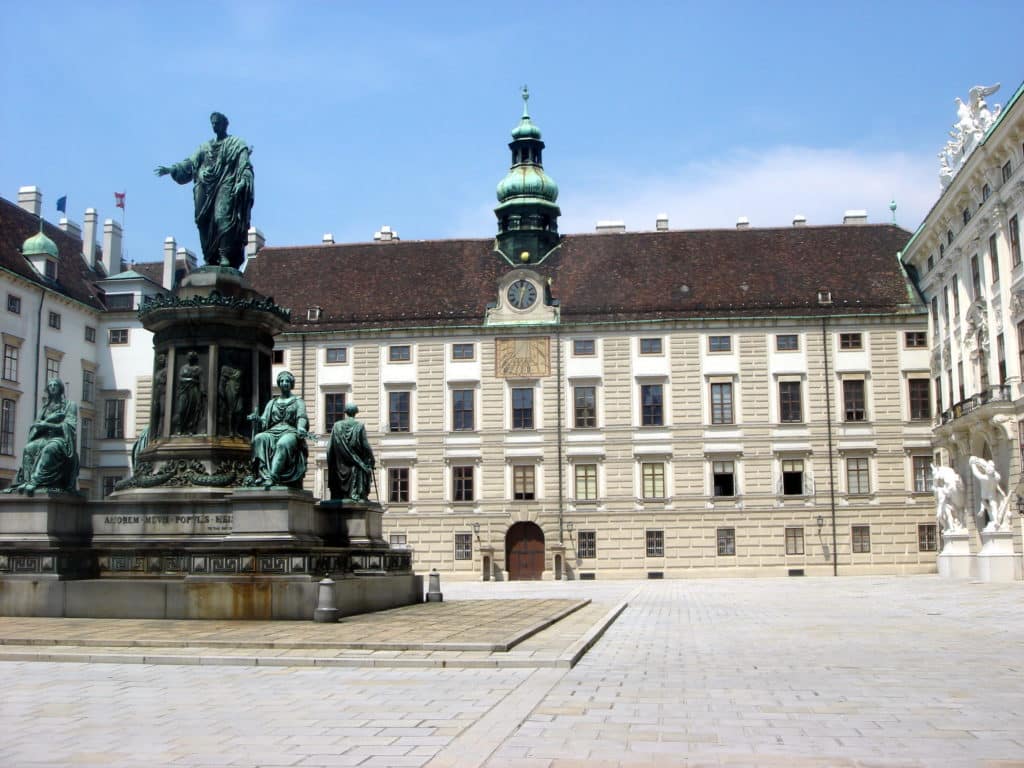 Uma parte de Hofburg vista de fora. Ele fica em uma praça histórica com uma grande estátua em frente a um grande edifício com uma torre de relógio. O céu está limpo e ensolarado.