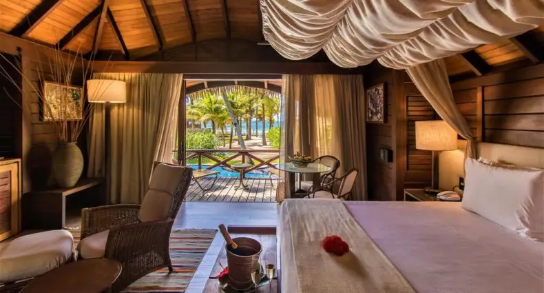 Quarto em bangalô de madeira com cama de dossel, área de estar, portas abertas para um deck e vista para as palmeiras e a água.