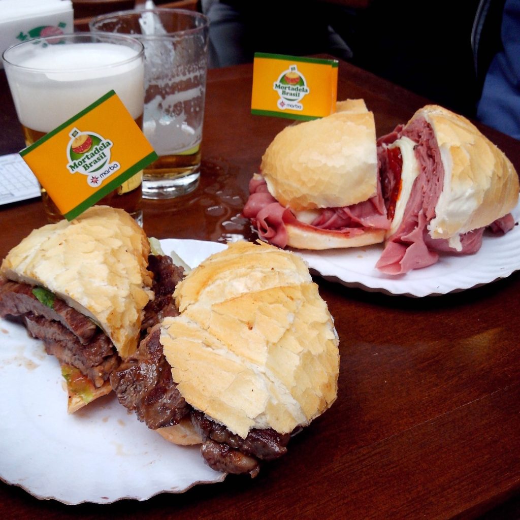 Na imagem vemos dois lanches em pão francês, um de carne e outro de mortadela, com dois copos de cerveja ao fundo. Foto de Fábio Martins Santos Marron via Pixabay