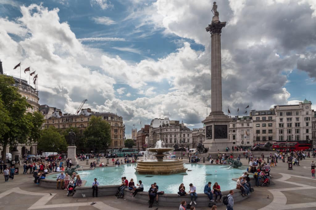 Vista da Trafalgar Square, com pessoas em volta da fonte e a coluna em homenagem ao Almirante Nelson ao fundo. Foto de Christian Reimer via Flickr.