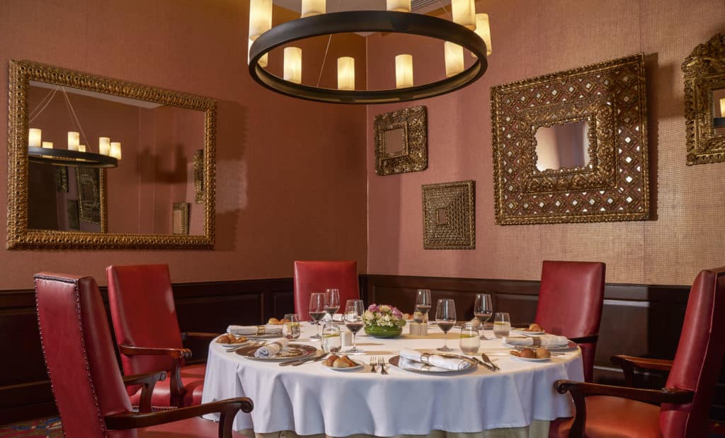 Foto com mesa redonda para jantar, cadeiras estofadas, e papel de parede rosé com espelhos em molduras douradas.