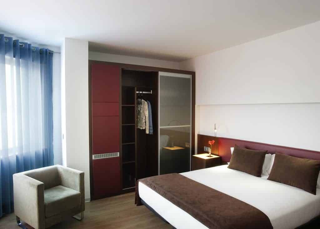 Quarto do Ayre Hotel Gran Vía, uma das recomendações de onde ficar em Barcelona. A cama tem lençol e travesseiros brancos e marrons, e há uma luminária em cima da mesinha de cabeceira de cada lado.
Há uma poltrona bege em frente da janela coberta por uma cortina azulada, e um guarda-roupa aberto está ao lado esquerdo.