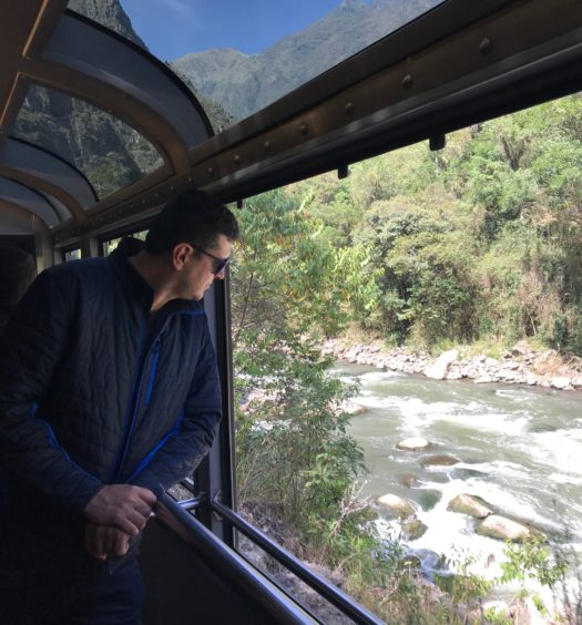 Foto de Flávio Antunes no trem aberto do vagão 360, com vista panorâmica da paisagem entre Cusco e Machu Picchu