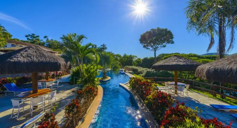 Uma piscina iluminada pelo sol com formato sinuoso, cercada por palmeiras, guarda-sóis de palha e espreguiçadeiras sob um céu azul claro.