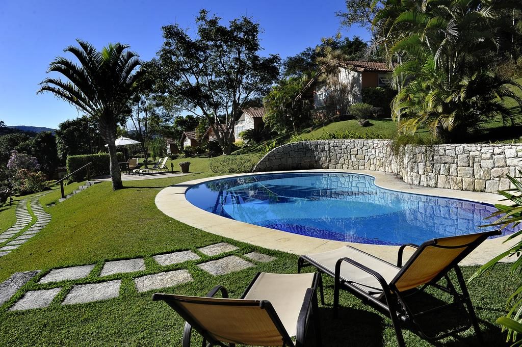 Pousada Recanto das Araras, área da piscina, com duas espreguiçadeiras em primeiro plano, a piscina ao centro, ao fundo a casa da pousada, localizada em volta de vasta vegetação, em um dia de céu azul.