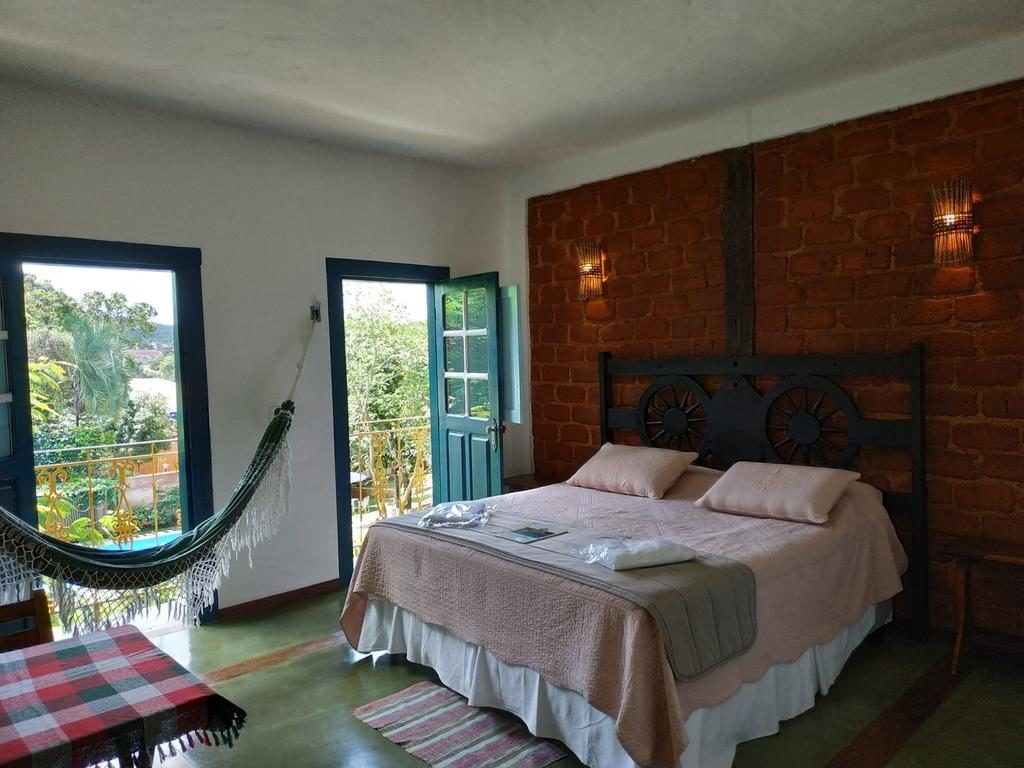 Quarto de casal da Pousada Villa Bia, cama com edredom rosa e travesseiros. sacada com vista para área externa da pousada e rede dentro do quarto.