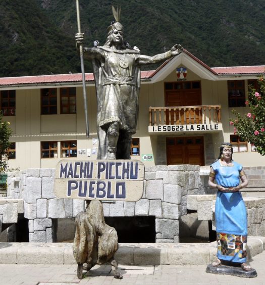 Estátuas no centro de Machu Picchu Pueblo com placa indicativa da cidade.