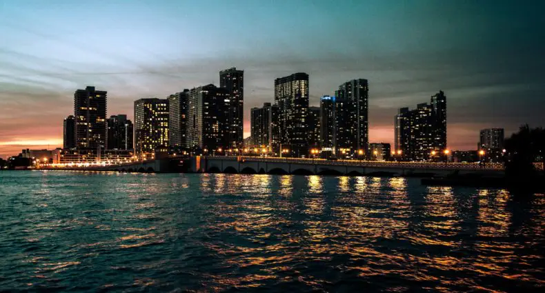 Horizonte de Miami ao anoitecer com edifícios iluminados refletindo na água, uma ponte visível em primeiro plano sob um céu escuro. Representa Onde ficar em Miami.