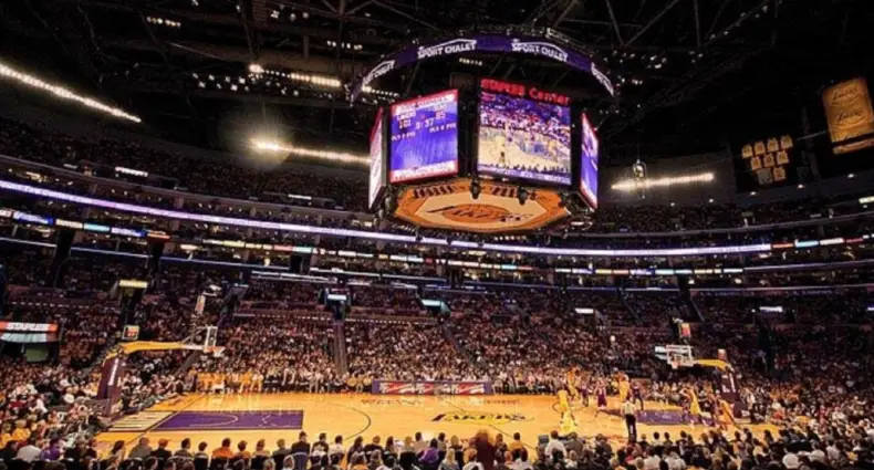 Um jogo de basquete está acontecendo em uma grande arena coberta, com um placar digital e um público lotado.