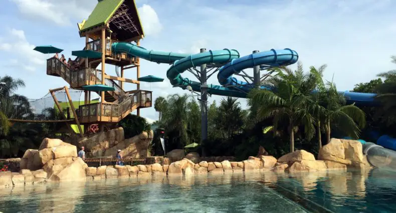 Uma grande estrutura de toboágua do O Dolphin Plunge, um dos principais tobogãs do Aquatica Orlando, com tubos azuis fica ao lado de uma piscina, cercada por pedras e palmeiras sob um céu azul.
