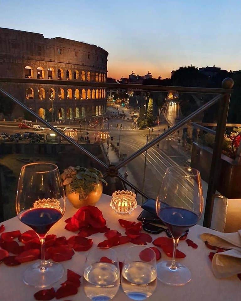 Jantar com luz baixa, taças de vinho e pétalas de rosa em mesa com vista direta para o Coliseu, uma boa pedida para lua de mel na Itália