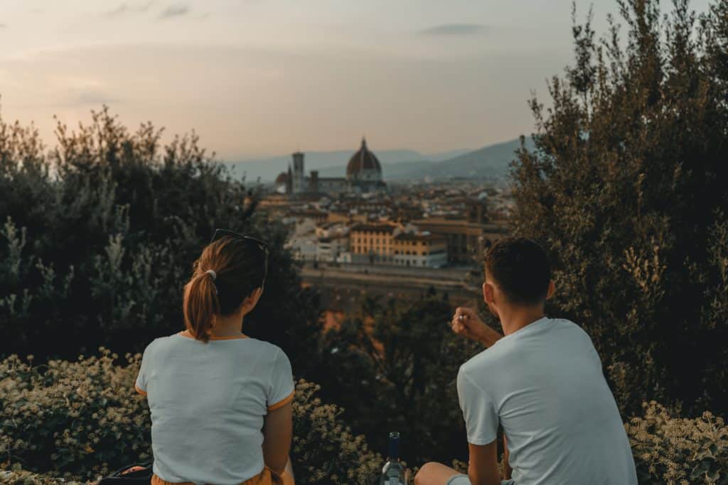 Casal com camisetas brancas sentados próximo à mata e observando a cidade de Florença ao fundo, uma das cidades indicadas para lua de mel na Itália