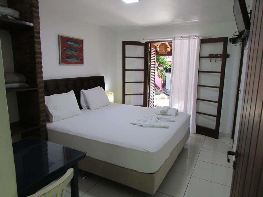 Uma cama de casal bem arrumada em um quarto bem iluminado na Pousada Bawa na Praia do Félix, com portas abertas que dão para um pátio; as toalhas estão dobradas sobre a cama.