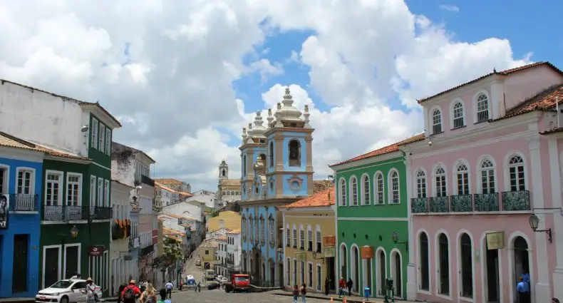 Uma rua de paralelepípedos repleta de edifícios coloridos leva a uma igreja barroca sob um céu nublado em Salvador, Brasil.