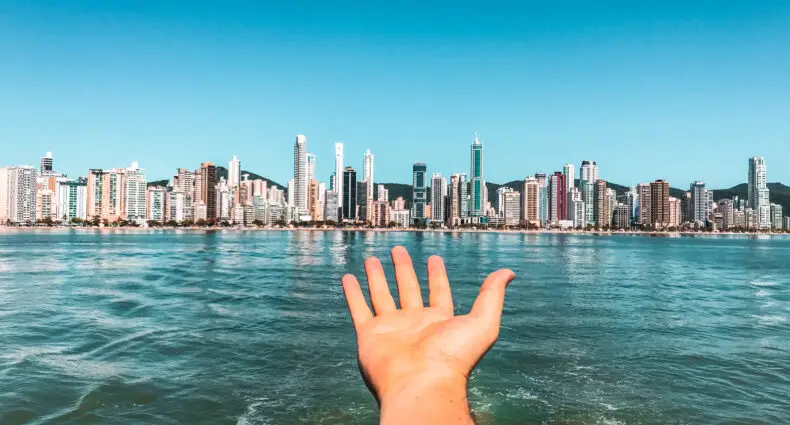 Vista do barco para a Praia Central e uma mão apresentando a paisagem. No fundo a areia da praia e os prédios. Imagem para ilustrar post sobre o que fazer em Balneário Camboriú.