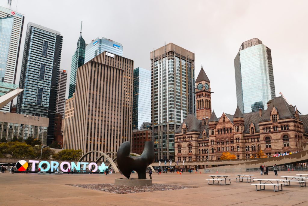 Praça de Toronto com escultura ao centro e placa com nome da cidade, além de prédios ao fundo