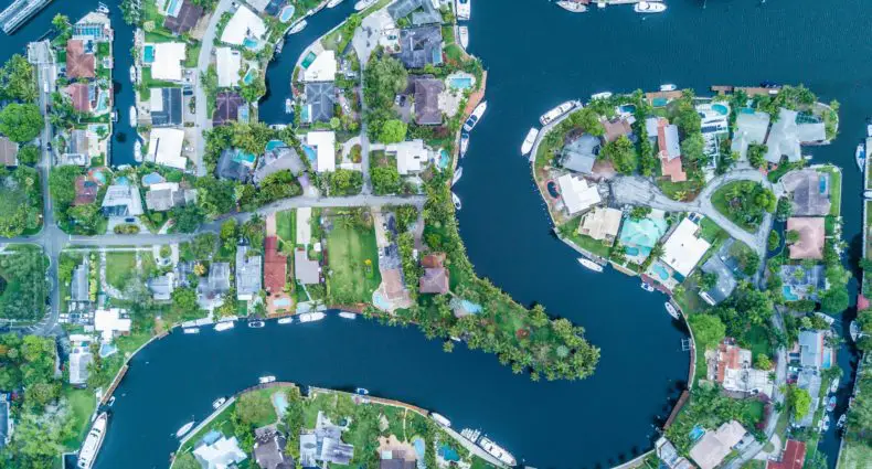 Vista aérea de um bairro residencial à beira de um canal com formato sinuoso, com casas cercadas por árvores, jardins e barcos ancorados nas margens da água, em região costeira ensolarada de Fort Lauderdale.