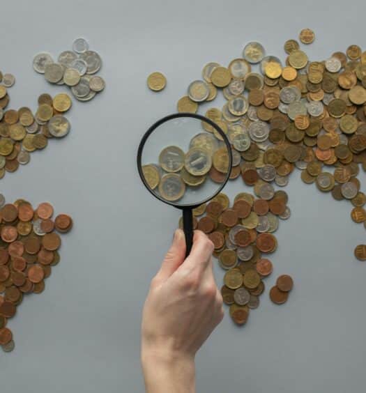 Mapa mundi desenhado com moedas enquanto uma mão segura uma lupa no centro da foto, para representar quanto custa atendimento médico no exterior