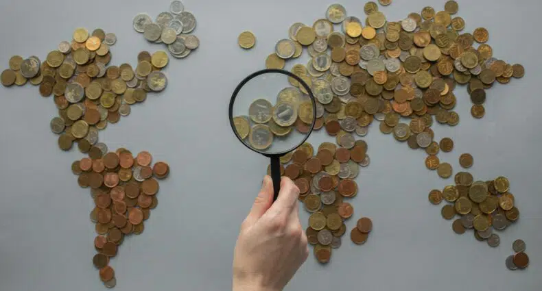 Mapa mundi desenhado com moedas enquanto uma mão segura uma lupa no centro da foto, para representar quanto custa atendimento médico no exterior