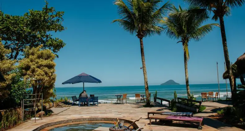 Área da piscina com espreguiçadeiras, palmeiras e mesas com vista para o oceano e uma ilha distante sob um céu azul claro.