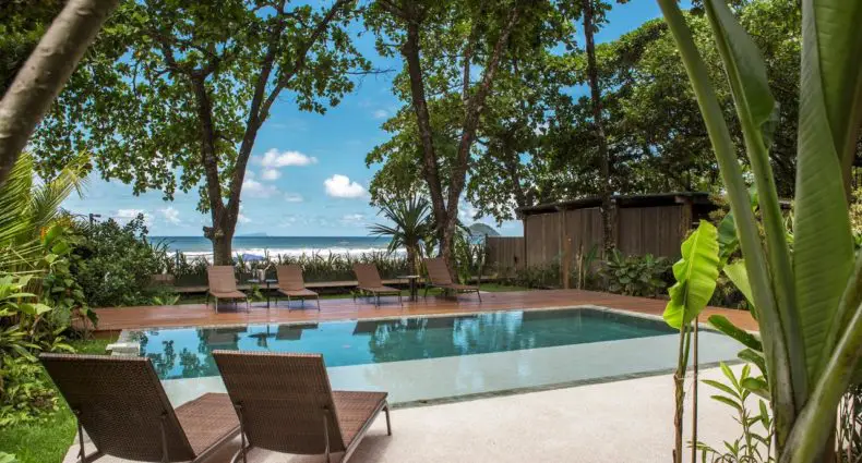 Piscina externa retangular do Flat Pitanga com espreguiçadeiras, cercada por árvores e plantas, com vista para o mar ao fundo.