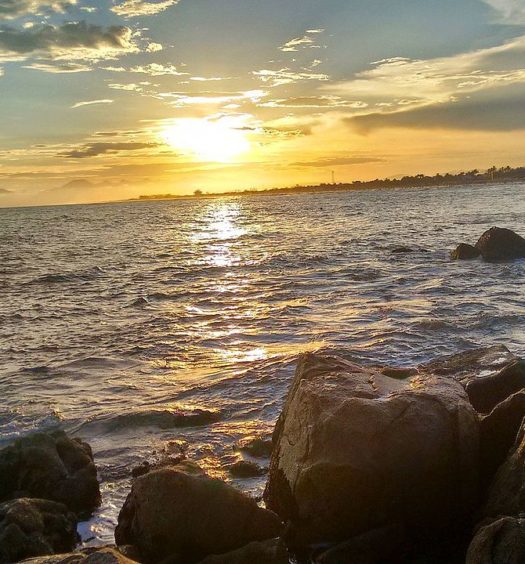 Vista de praia em Itanhaém, com rochas na beira das águas, durante o fim da tarde
