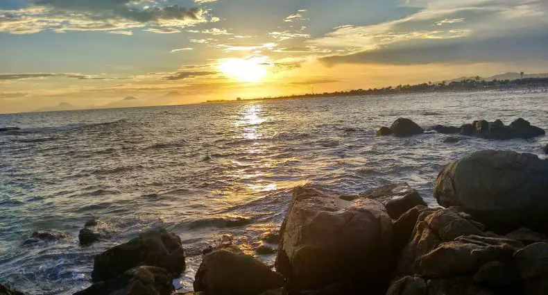 Vista de praia em Itanhaém, com rochas na beira das águas, durante o fim da tarde