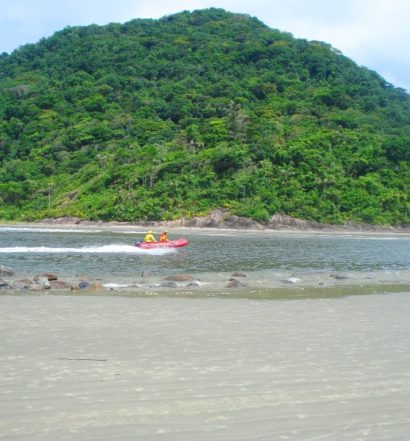 Mata em ilha, águas com pessoas passando em barco, e faixa de areia da Praia de Guaraú, em Peruíbe