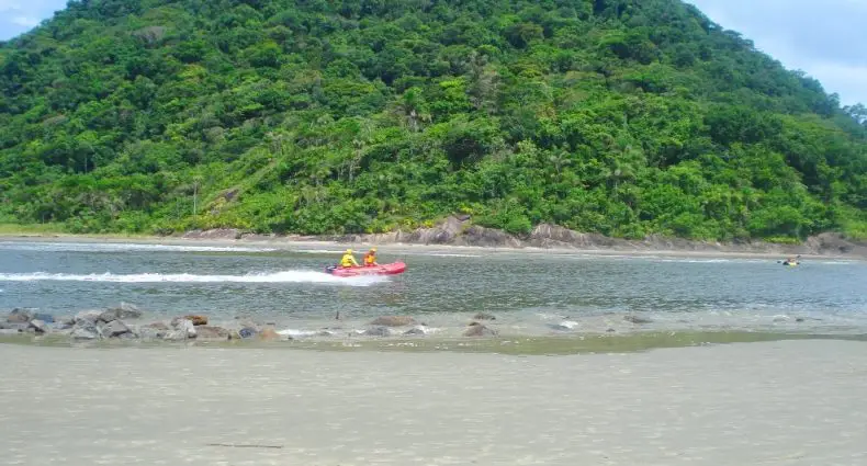 Mata em ilha, águas com pessoas passando em barco, e faixa de areia da Praia de Guaraú, em Peruíbe