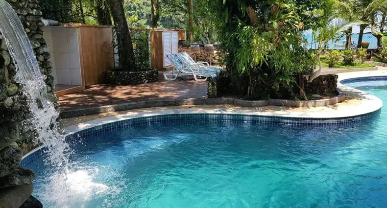 Piscina curva da Pousada Morada das Ilhas, uma das pousadas em Boiçucanga com água azul clara, cachoeira de pedra, espreguiçadeiras e árvores que proporcionam sombra.