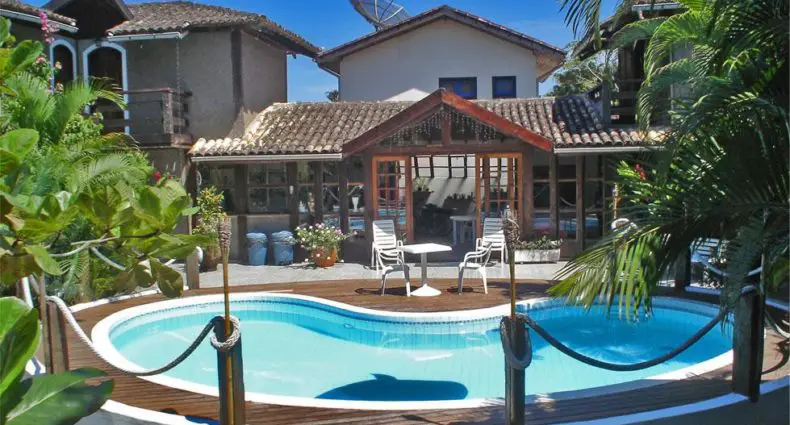Uma pequena piscina redonda com corrimão de corda fica em frente a uma casa com telhado de telha e palmeiras, na propriedade da A Pousada Praia da Baleia, para representar pousadas na Praia da Baleia.