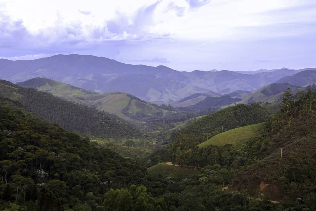 Uma paisagem montanhosa com colinas e vales verdes sob um céu nublado, oferecendo uma vista panorâmica de picos distantes.
