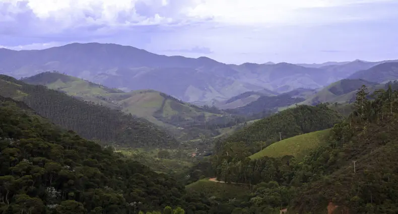 Uma paisagem montanhosa com colinas e vales verdes sob um céu nublado, oferecendo uma vista panorâmica de picos distantes.