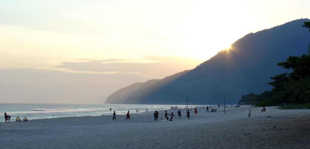 Faixa de areia em Maresias, São Sebastião. Alguas pessoas estão andando, jogando e olhando o mar, que está do lado esquerdo.