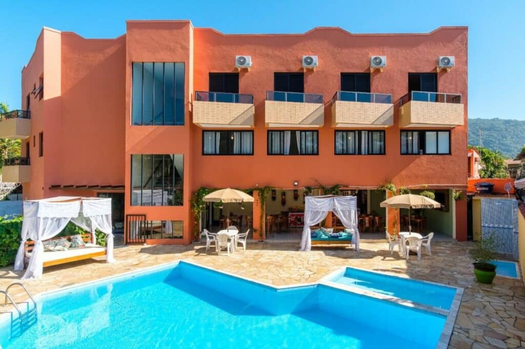 Vista externa do Hotel Ponta das Toninhas, com paredes laranja, sacada com ar-condicionado nos quartos, e, no pátio térreo, piscina, pergolados, mesas, cadeiras e guarda-sóis