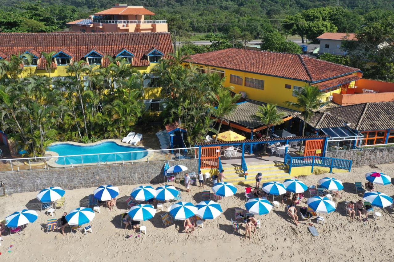 Vista aérea da Kaliman Pousada à beira-mar com piscina, palmeiras e pessoas sob guarda-sóis azuis e brancos na areia da praia.