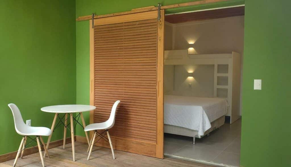 Quarto na Pousada Vila Jacaa, uma sacada com porta de madeira, um quarto branco com móveis também brancos, uma cama de casal e uma beliche, na varanda, uma mesa com duas cadeiras